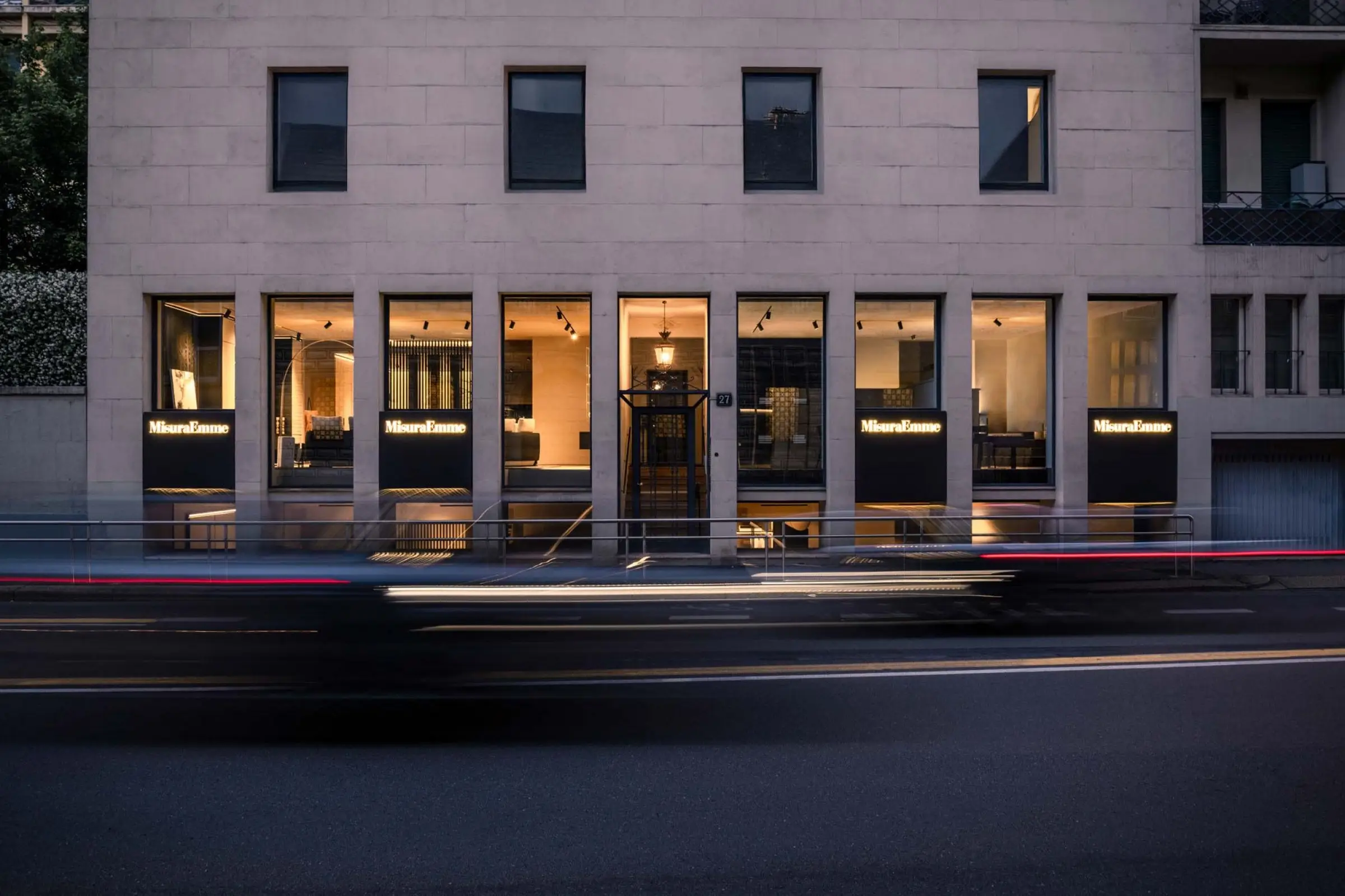 La facciata del flagship store MisuraEmme in via Uberto Visconti di Modrone a Milano, al tramonto, con le vetrine accese e le scie delle auto